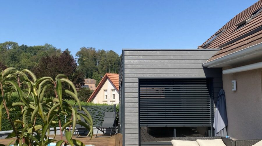 Vue d'ensemble d'un agrandissement de maison moderne réalisé en ossature bois avec bardage gris foncé, intégrant une piscine et une terrasse.