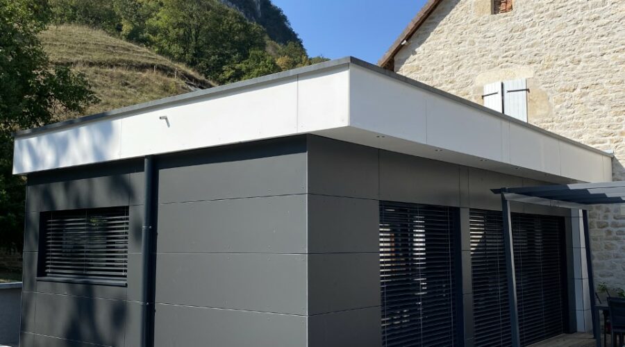 Extension de maison contemporaine en bois avec bardage gris foncé, grandes ouvertures et toit plat, attenante à une maison en pierre et ouvrant sur une terrasse en bois.