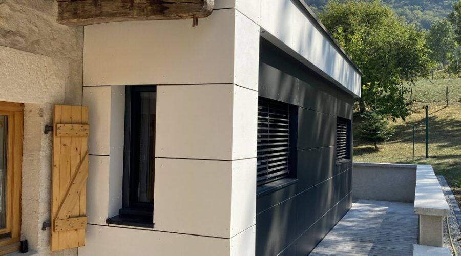 Détail d'une extension de maison en ossature bois avec un mélange de bardage sombre et clair, et ses fenêtres modernes, accolée à une maison en pierre.