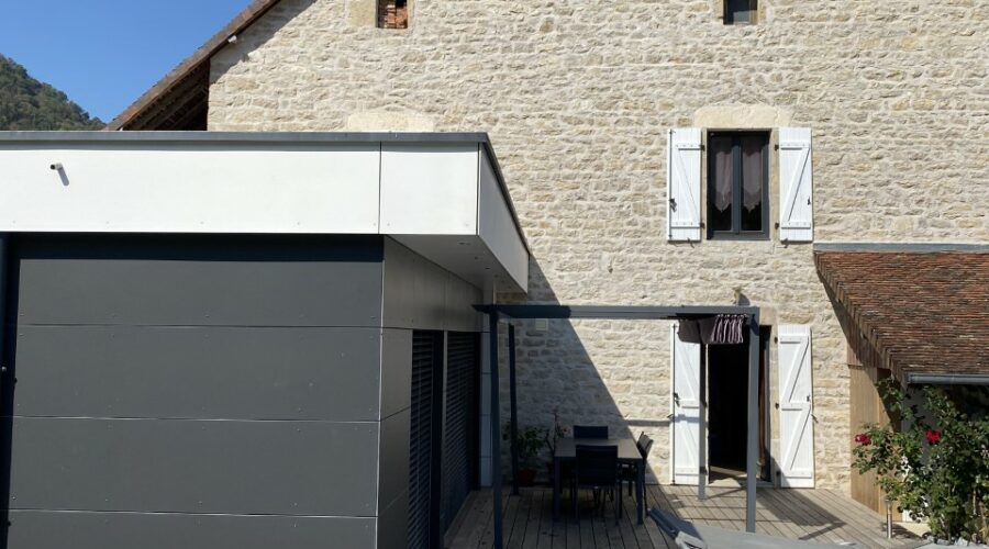 Agrandissement de maison en ossature bois, montrant le bardage moderne gris foncé, la vaste terrasse en bois et la façade en pierre de la maison principale.