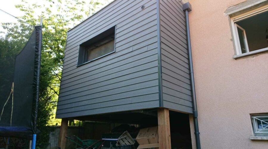 Vue arrière d'une extension de maison en ossature bois avec bardage gris foncé, montrant le toit plat et les ouvertures.
