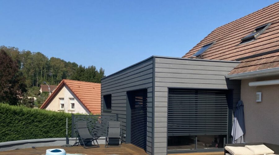 Extension moderne en ossature bois avec bardage gris foncé, située près d'une piscine et d'une terrasse en bois, avec brise-soleil orientables (BSO).