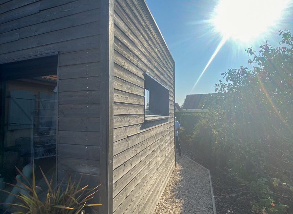 Façade latérale d'une extension en ossature bois avec bardage foncé, dotée d'une fenêtre verticale et d'un chemin de graviers, baignée de soleil.