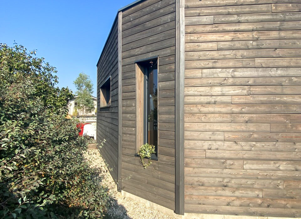 Gros plan sur le bardage en mélèze foncé d'une extension en bois, avec une petite fenêtre verticale intégrée, et un chemin de graviers.