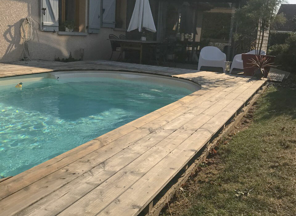Vue d'une extension en ossature bois sombre s'intégrant à une maison existante, avec une piscine et une terrasse en bois clair en premier plan.