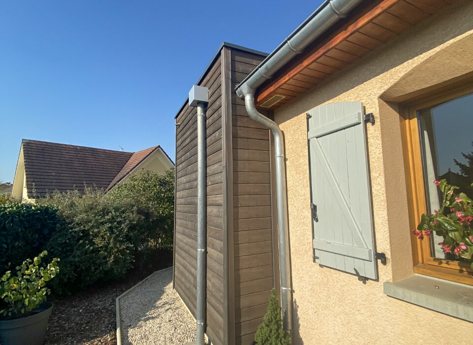 Vue latérale d'une extension en bois foncé accolée à une maison traditionnelle, montrant le raccordement discret de la gouttière et l'harmonie des matériaux.
