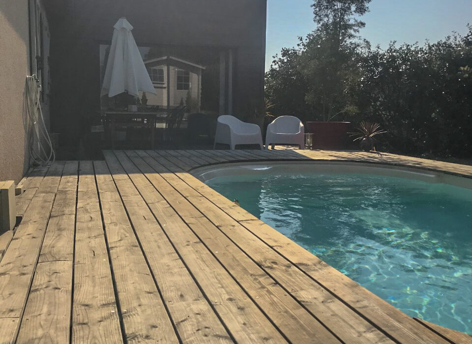 Perspective d'une terrasse en bois entourant une piscine, avec un espace de vie extérieur ombragé par une extension moderne en arrière-plan.