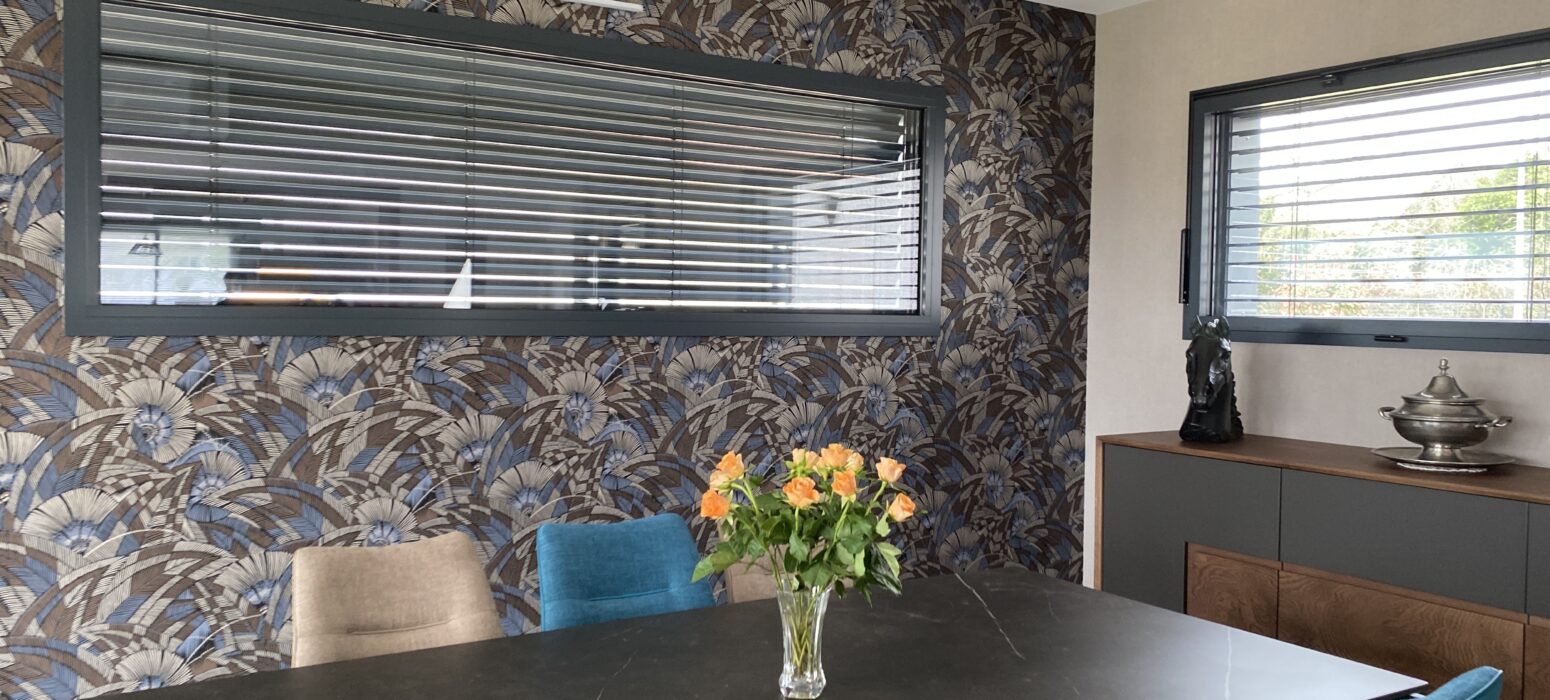 Salle à manger moderne dans une extension en bois, avec table noire, chaises colorées, grand papier peint à motifs floraux et fenêtre horizontale équipée de brise-soleil orientables.