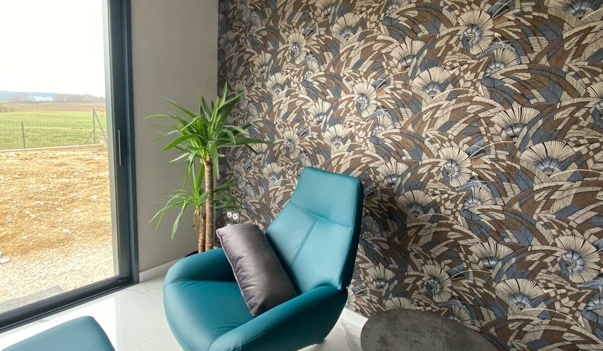Coin salon lumineux dans une extension bois, avec fauteuil design turquoise, table basse, mur décoré d'un papier peint à motifs, et vue sur la nature extérieure.