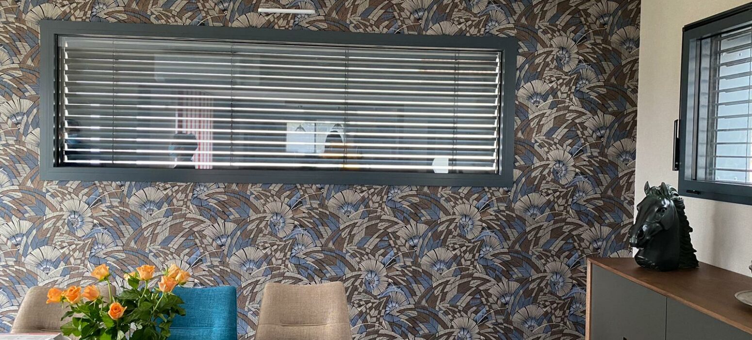 Détail de l'espace salle à manger d'une extension moderne, mettant en valeur le papier peint à motifs, le buffet bas et la fenêtre avec ses brise-soleil intégrés.