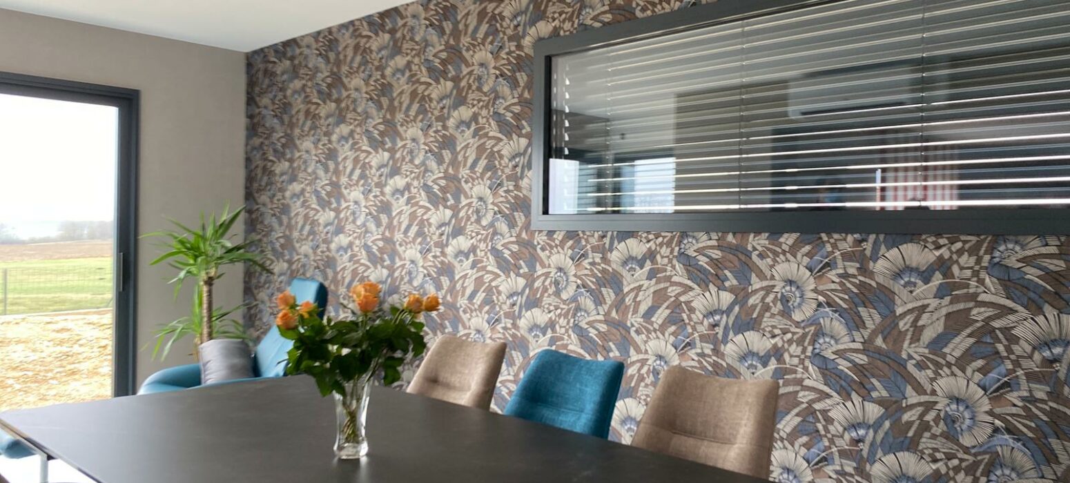 Salle à manger dans une extension bois, avec table élégante, chaises aux couleurs variées, papier peint floral vibrant et large fenêtre pour un apport de lumière optimal.